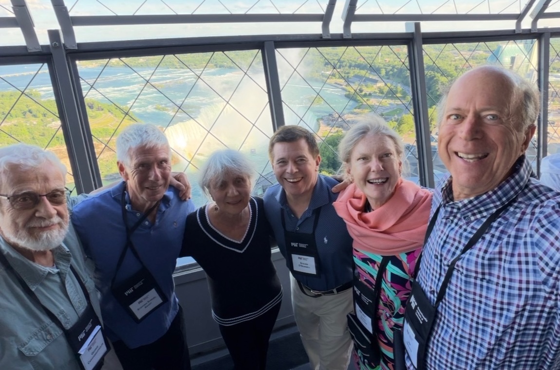 George Carayannopoulos, Stephen Abany, Margaret Carayannopoulos, Steve O'Donnell, and Coral Sullivan smile as Jim Bricker takes a selfie of the group in front of a large window overlooking Niagara Falls