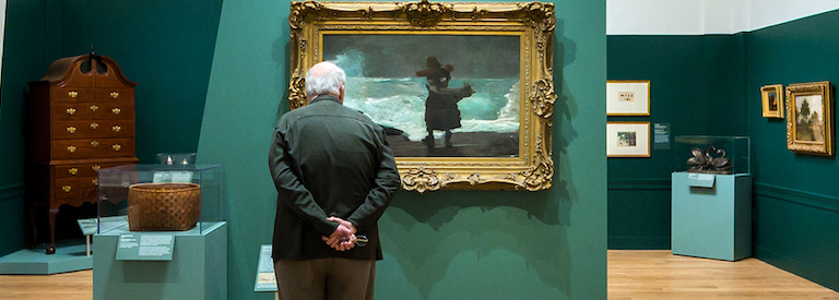 An older gentleman in a deep teal gallery room regards a painting of a woman on the seashore.